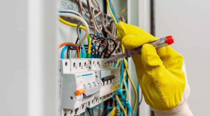An electrician wearing gloves uses a tester to inspect and maintain an electrical panel with multiple colored wires.