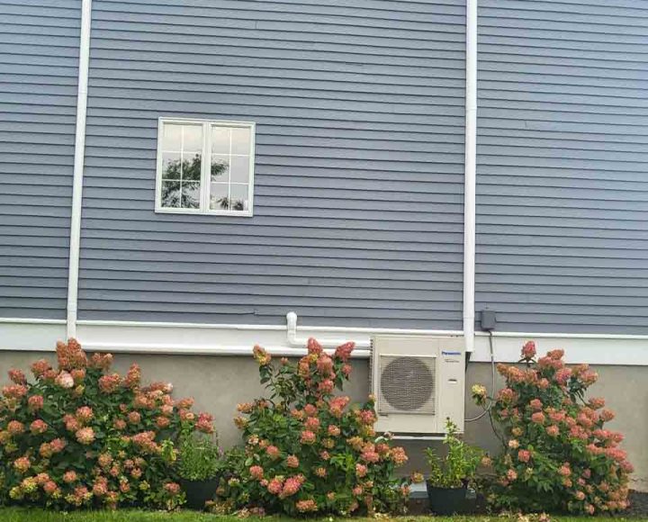 A Panasonic air conditioning unit installed on the exterior of a blue house, surrounded by vibrant flowering shrubs.