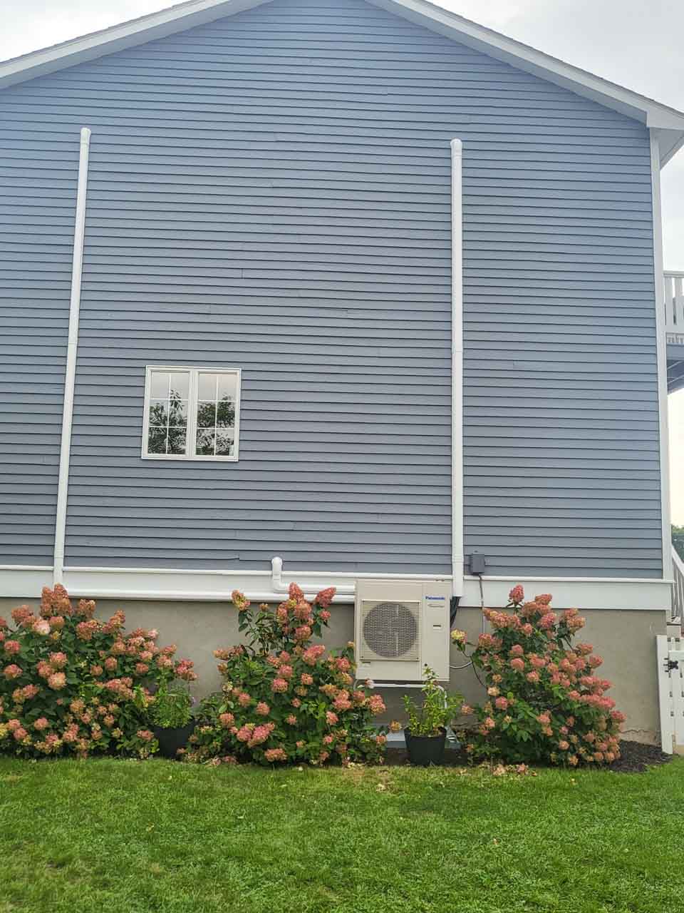 An air conditioning unit installed on the exterior wall of a blue house, surrounded by colorful flowers and greenery.