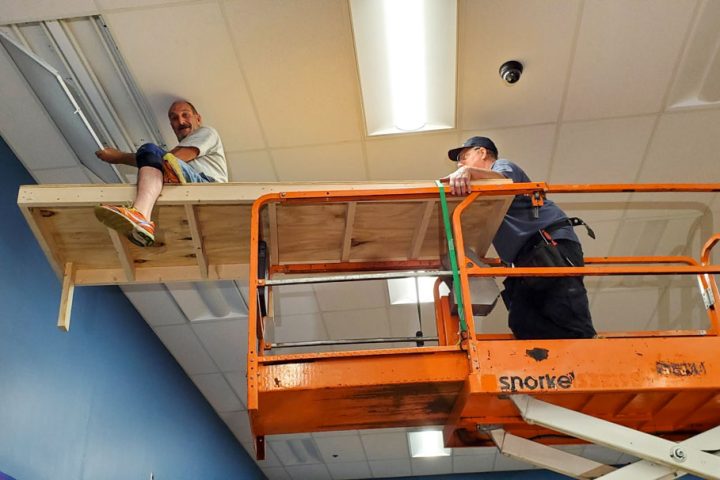 Two workers on an orange lift platform collaborate to install or repair ceiling fixtures in a commercial space.