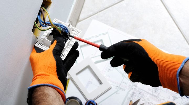A professional electrician installs and tests an electrical outlet using tools and safety gear.