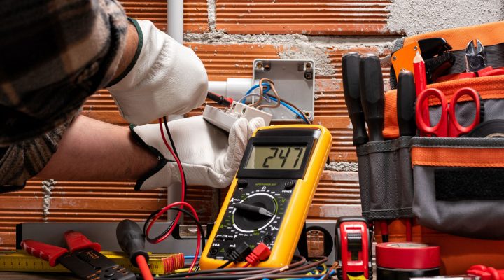 An electrician wearing gloves uses a multimeter to test electrical connections during a pre-inspection.